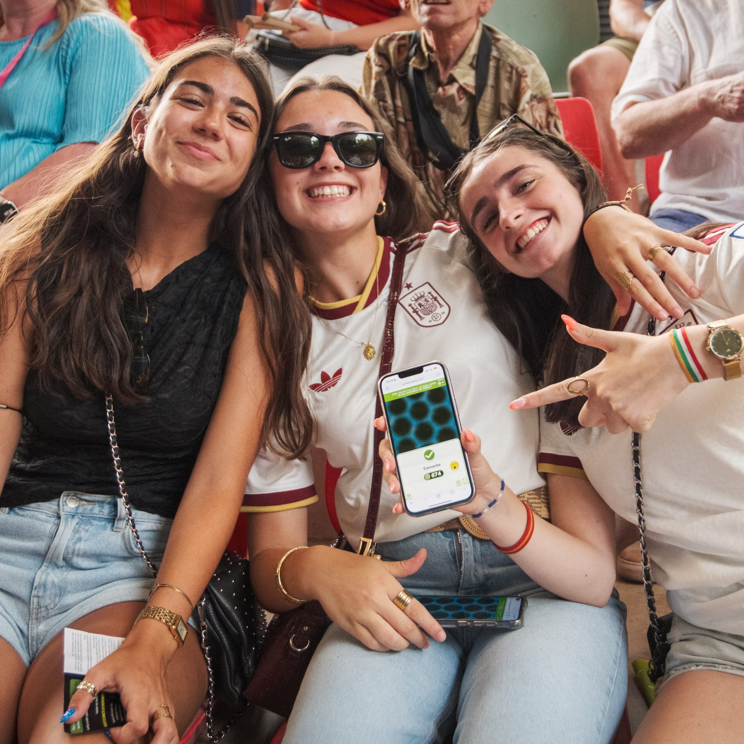 Tres jóvenes con camisetas de la selección española sonríen a cámara; una sostiene el móvil con una Partida Multijugador de Educaplay marcando «Correcto».