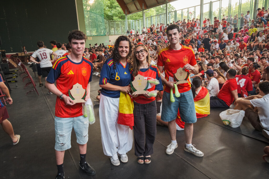 Four participants smile as they pose with numbered wooden Educaplay trophies and neck fans, with the court crowd in the background.